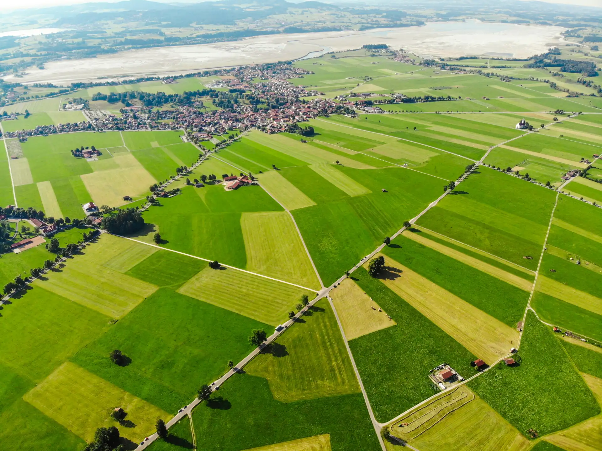 Luftbild grüner Wiesenparzellen mit Dorf und Seeufer im Alpenvorland